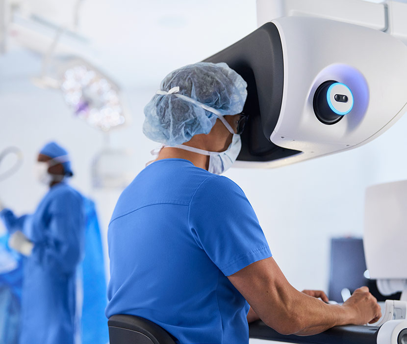 Person sits at console while controlling a robot in an operating room.